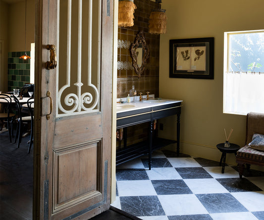 An open rustic wooden door with white decorative metal scrollwork reveals an interior room with a black-and-white checkered floor, a dark vanity table, warm yellow walls, and hanging woven pendant lights.