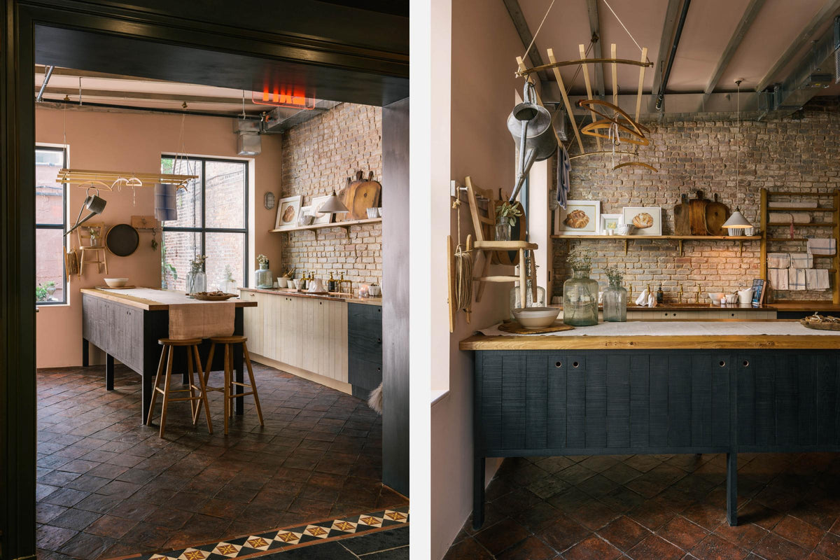 A calm and simple Sebastian Cox kitchen features a long sink run with two sets of copper sinks and aged brass Mayan taps, a copper worktop, a shelf, and a prep table with a solid oak worktop. Adorned with vintage items, artisan ceramics, wooden chopping boards, framed illustrations, tea towels, and wildflowers in large glass containers, this kitchen creates a chic New York industrial space that celebrates a love of natural materials.