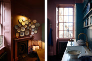 Light streams through sash windows into a moody room with original Georgian features, dusky wine-coloured walls, and dark teal Shaker cabinetry. The display includes a sink run with a fluted ceramic sink, aged brass taps, classic brass door hardware, a white Carrara marble worktop, a tall countertop cupboard, tongue-and-groove wall panelling, and shelves showcasing a selection of classic decorative plates.