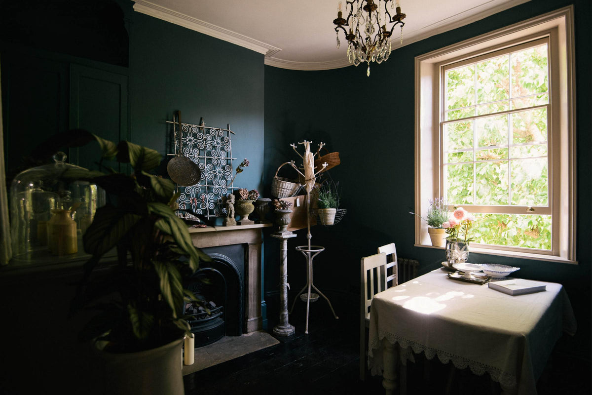 A dramatic image of a spacious, muted-blue dining area, beautifully decorated with antique furnitures, vintage ceramics, and luscious green houseplants.