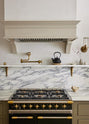 A closer look at an elegant mushroom-brown, classic English kitchen features a cooker run with an Arabescato marble worktop, splashback, and shelf. Beautifully decorated with artisan ceramics, brass classic shelf brackets, a deVOL aged brass pot filler, and a cast-iron teapot, it adds a sophisticated touch to the elegant kitchen.
