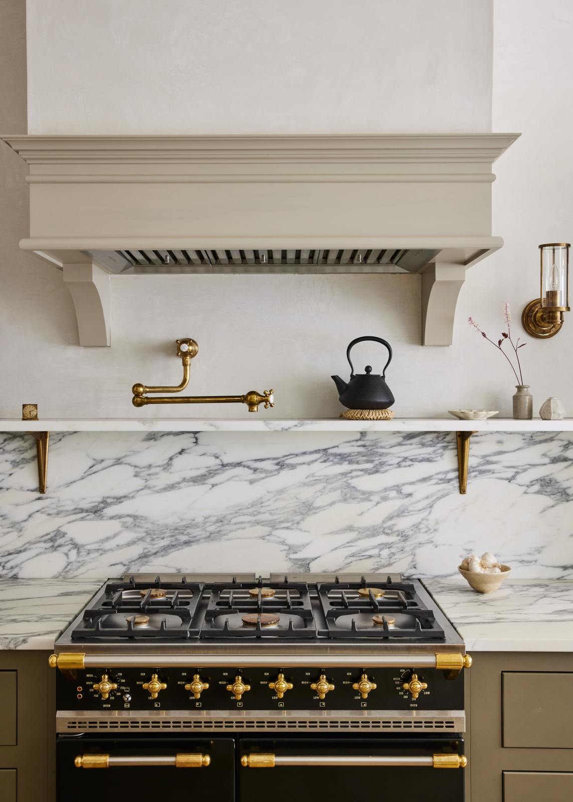A closer look at an elegant mushroom-brown, classic English kitchen features a cooker run with an Arabescato marble worktop, splashback, and shelf. Beautifully decorated with artisan ceramics, brass classic shelf brackets, a deVOL aged brass pot filler, and a cast-iron teapot, it adds a sophisticated touch to the elegant kitchen.
