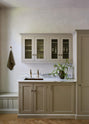 An elegant mushroom-brown, classic English kitchen features a wet bar with a single copper sink, aged brass Mayan taps, an Arabescato marble worktop, and reeded wall cupboards. Beautifully decorated with artisan ceramic and green bell flowers, it adds a sophisticated touch to the elegant kitchen.