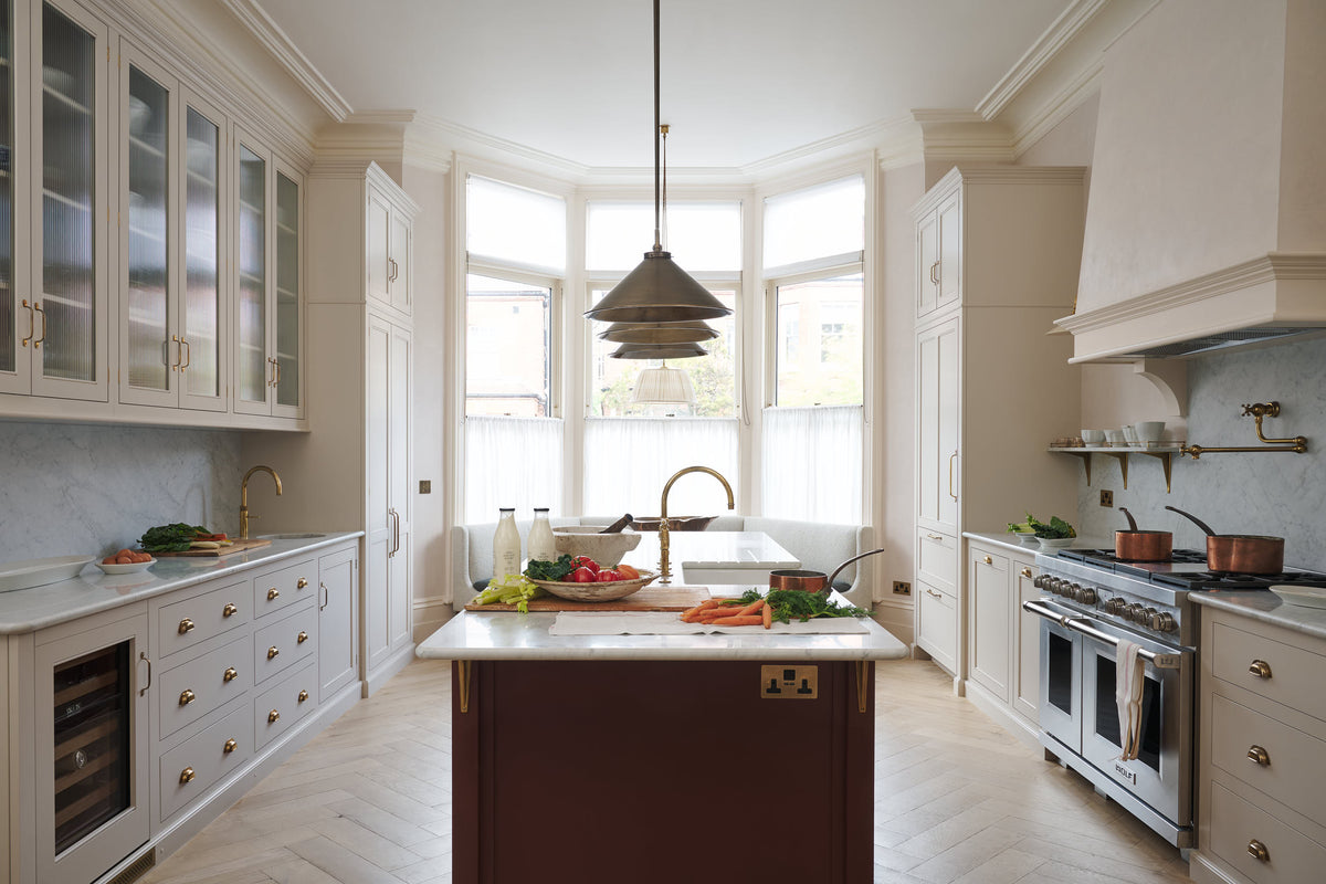 A grand yet understated Classic English kitchen, this space features a sink run with reeded glazed wall cupboards, full-height cabinetry, a dedicated cooker run with a canopy, and an island with a Carrara marble sink. Styled with vintage French copper pans, artisan ceramics, and baskets of fresh vegetables and eggs, it feels wonderfully light, airy, and perfectly suited to relaxed, sociable living.