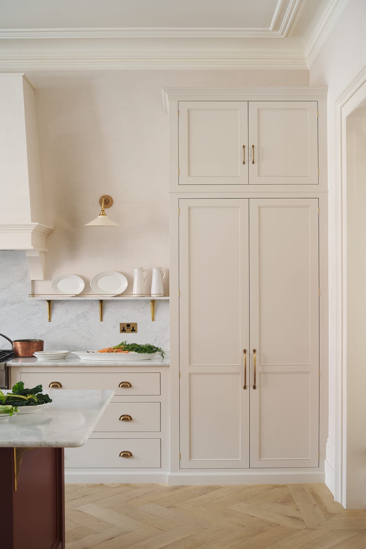 A quiet corner of a grand yet understated Classic English kitchen features a dedicated cooker run with a canopy, a full-height cupboard, and an island. Lovingly adorned with vintage French copper pans, artisan ceramics, and fresh vegetables, it is perfectly suited to relaxed, sociable living.