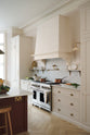 An oblique view of a grand yet understated Classic English kitchen features a dedicated cooker run with a canopy, a full-height cupboard, and an island. Lovingly adorned with vintage French copper pans, artisan ceramics, and fresh vegetables, it is perfectly suited to relaxed, sociable living.