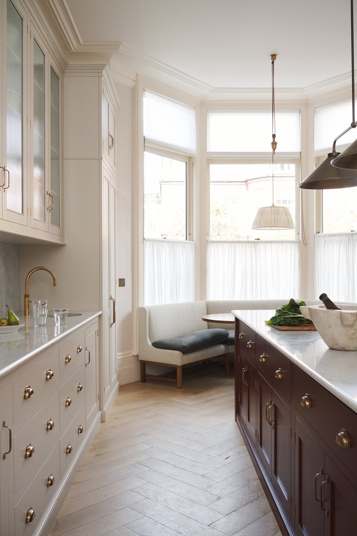 A grand yet understated Classic English kitchen, this space features a sink run with reeded glazed wall cupboards, full-height cabinetry, and an island with a Carrara marble sink. Styled with vintage French copper pans, artisan ceramics, and baskets of fresh fruits and vegetables, it feels wonderfully light, airy, and perfectly suited to relaxed, sociable living.