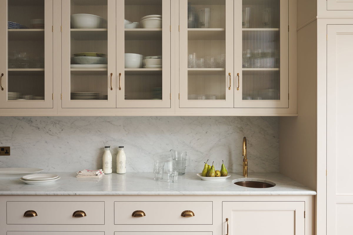 A grand yet understated Classic English kitchen, this space features a sink run with reeded glazed wall cupboards and full-height cabinetry. Styled with artisan ceramics and a bowl of fresh green pears, it feels wonderfully light, airy, and perfectly suited to relaxed, sociable living.