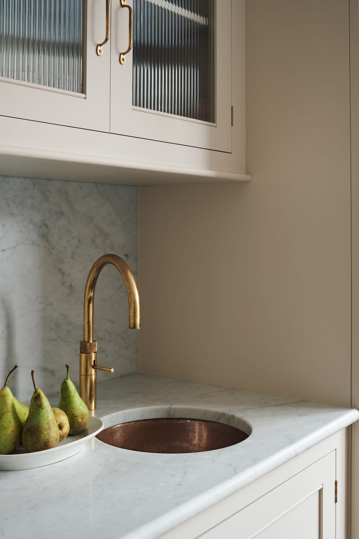 A closer look at the grand yet understated Classic English kitchen reveals a sink run with reeded glazed wall cupboards, full-height cabinetry, and an aged copper sink. Styled with artisan ceramics, and a bowl of fresh, perfectly suited to relaxed, sociable living.