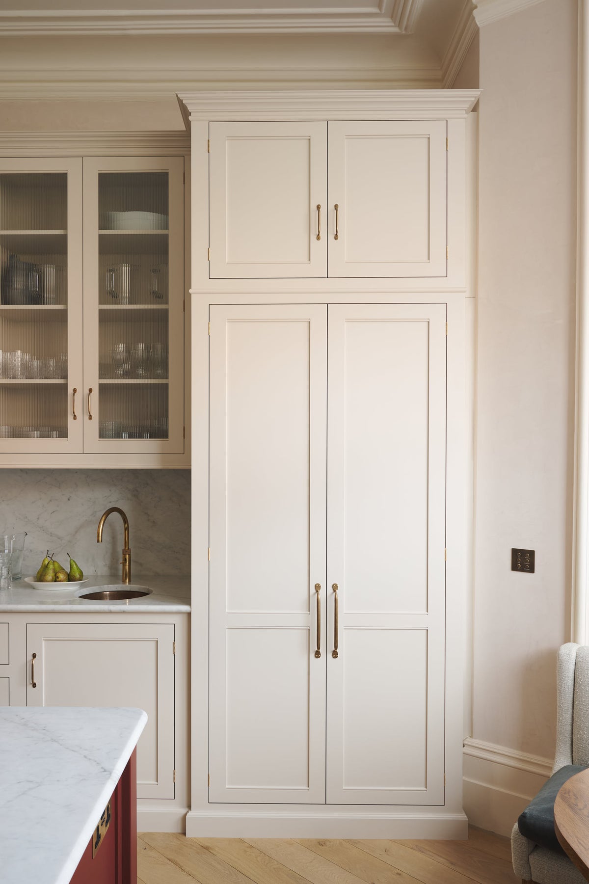 A quiet corner of a grand yet understated Classic English kitchen, this space features a sink run with reeded glazed wall cupboards, full-height, and aged copper cabinetry. Styled with artisan ceramics and a bowl of fresh green pears, it feels wonderfully light, airy, and perfectly suited to relaxed, sociable living.