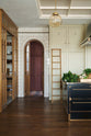One side of a grand room—with a high panelled ceiling, glossy white tiled walls, and curved archways flanking a huge bank of creamy mushroom full-height classic cupboards with a polished oak ladder—features a black kitchen island with gold accents and a canopy. Beyond, an arched doorway leads to a refectory-red room housing the large haberdasher’s pantry.