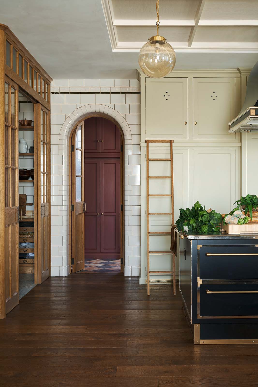 One side of a grand room—with a high panelled ceiling, glossy white tiled walls, and curved archways flanking a huge bank of creamy mushroom full-height classic cupboards with a polished oak ladder—features a black kitchen island with gold accents and a canopy. Beyond, an arched doorway leads to a refectory-red room housing the large haberdasher’s pantry.