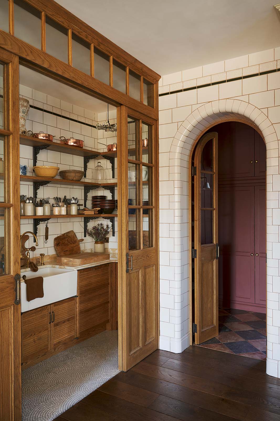 One side of a grand room with a high panelled ceiling, traditional glossy white tiled walls with curved archways. A large haberdasher’s pantry complete with its own Haberdasher’s sink run, topped with a marble worktop, large shelves, and an assorted collection of ceramic plates, bowls, pots, pans, and silverware.