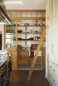 Looking down the grand kitchen with a high panelled ceiling, traditional glossy white tiled walls, and a huge bank of creamy mushroom full-height classic cupboards with a polished oak ladder is a large haberdasher’s pantry. It’s complete with its own haberdasher’s sink run topped with a marble worktop, large shelves, and an assorted collection of ceramic plates, bowls, pots, pans, and silverware.