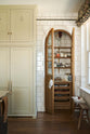 A small haberdasher’s pantry with a tiled interior and exterior sits just beyond a curved archway door, beside a grand wall of creamy mushroom full-height classic cupboards. The pantry features three shelves, a haberdasher’s open base cabinet with a marble worktop, and stores colourful dried ingredients in glass jars, along with fresh vegetables and spices.