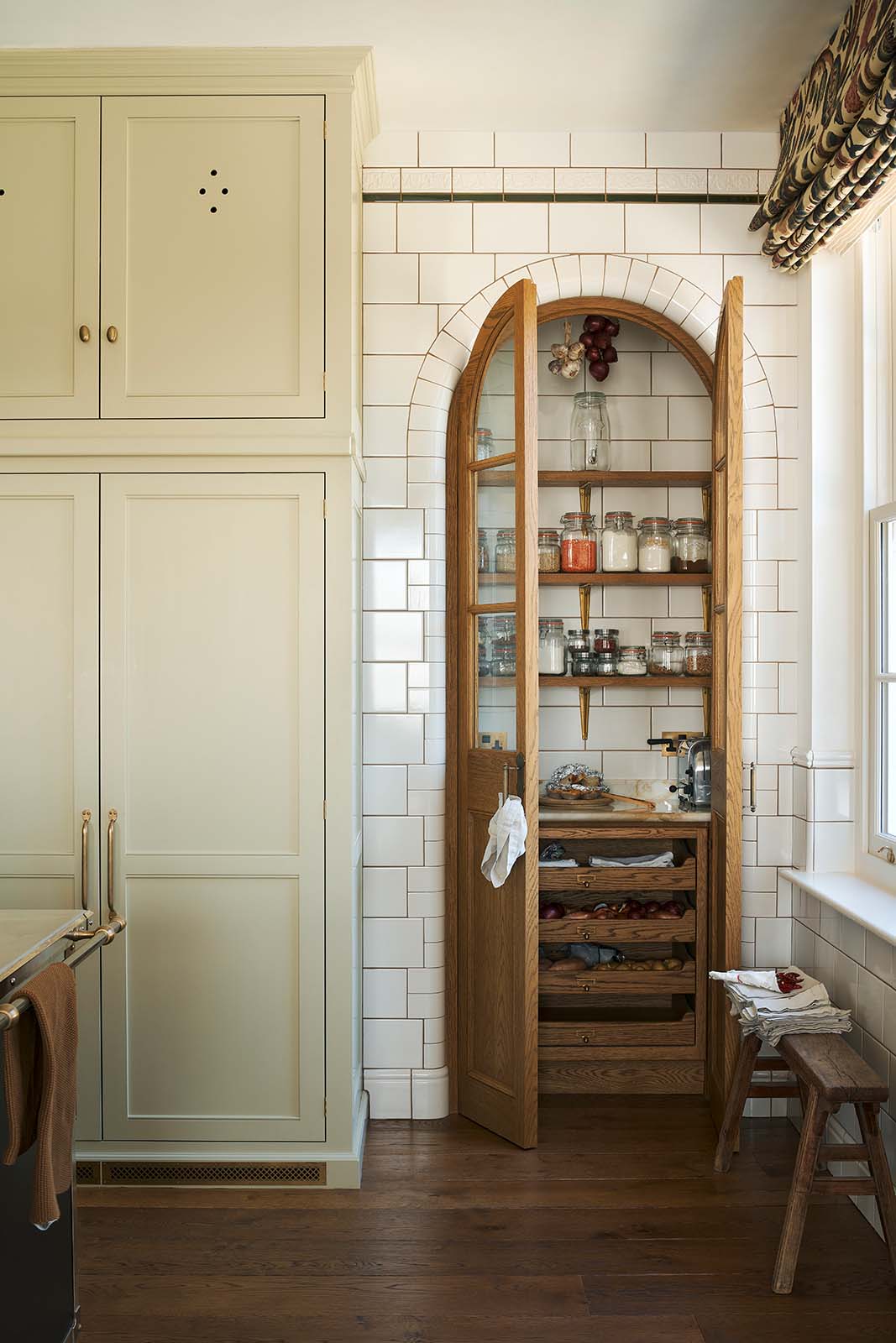 A small haberdasher’s pantry with a tiled interior and exterior sits just beyond a curved archway door, beside a grand wall of creamy mushroom full-height classic cupboards. The pantry features three shelves, a haberdasher’s open base cabinet with a marble worktop, and stores colourful dried ingredients in glass jars, along with fresh vegetables and spices.