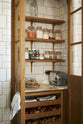 A closer, angled view reveals a small haberdasher’s pantry with tiled interior and exterior, nestled just beyond a curved archway door beside a grand wall of creamy mushroom full-height classic cupboards. The pantry features three shelves, an open base cabinet with a marble worktop, and stores colourful dried ingredients in glass jars, along with fresh vegetables and spices.