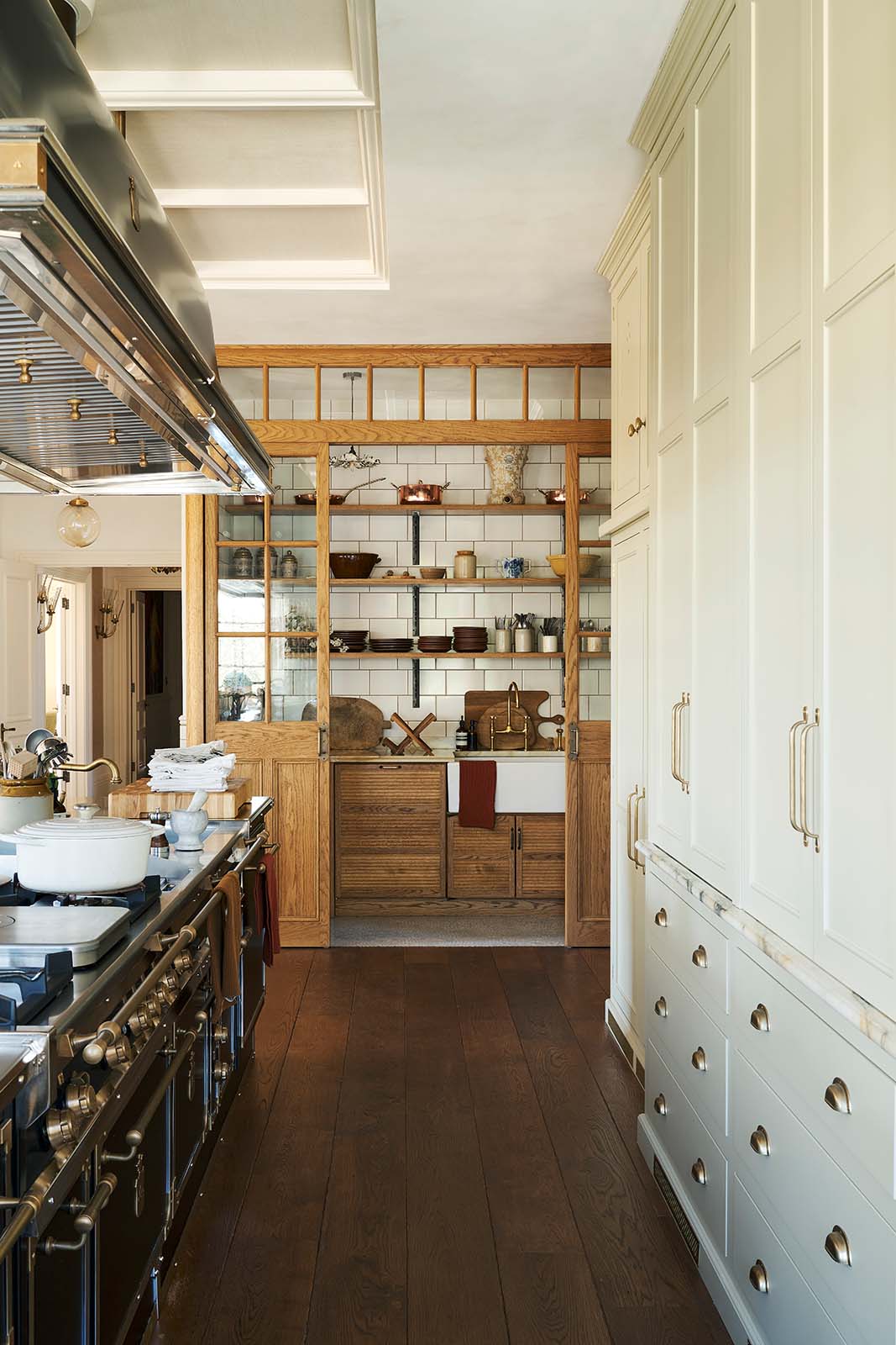 Looking down the grand kitchen with a high panelled ceiling, traditional glossy white tiled walls, and a huge bank of creamy mushroom full-height classic cupboards is a large haberdasher’s pantry. It’s complete with its own haberdasher’s sink run topped with a marble worktop, large shelves, and an assorted collection of ceramic plates, bowls, pots, pans, and silverware.