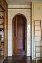 One side of a grand room—with a high panelled ceiling, glossy white tiled walls, and curved archways flanking a huge bank of creamy mushroom full-height classic cupboards with a polished oak ladder—features a black kitchen island with gold accents and a canopy. Beyond, an arched doorway leads to a refectory-red room housing the large haberdasher’s pantry.