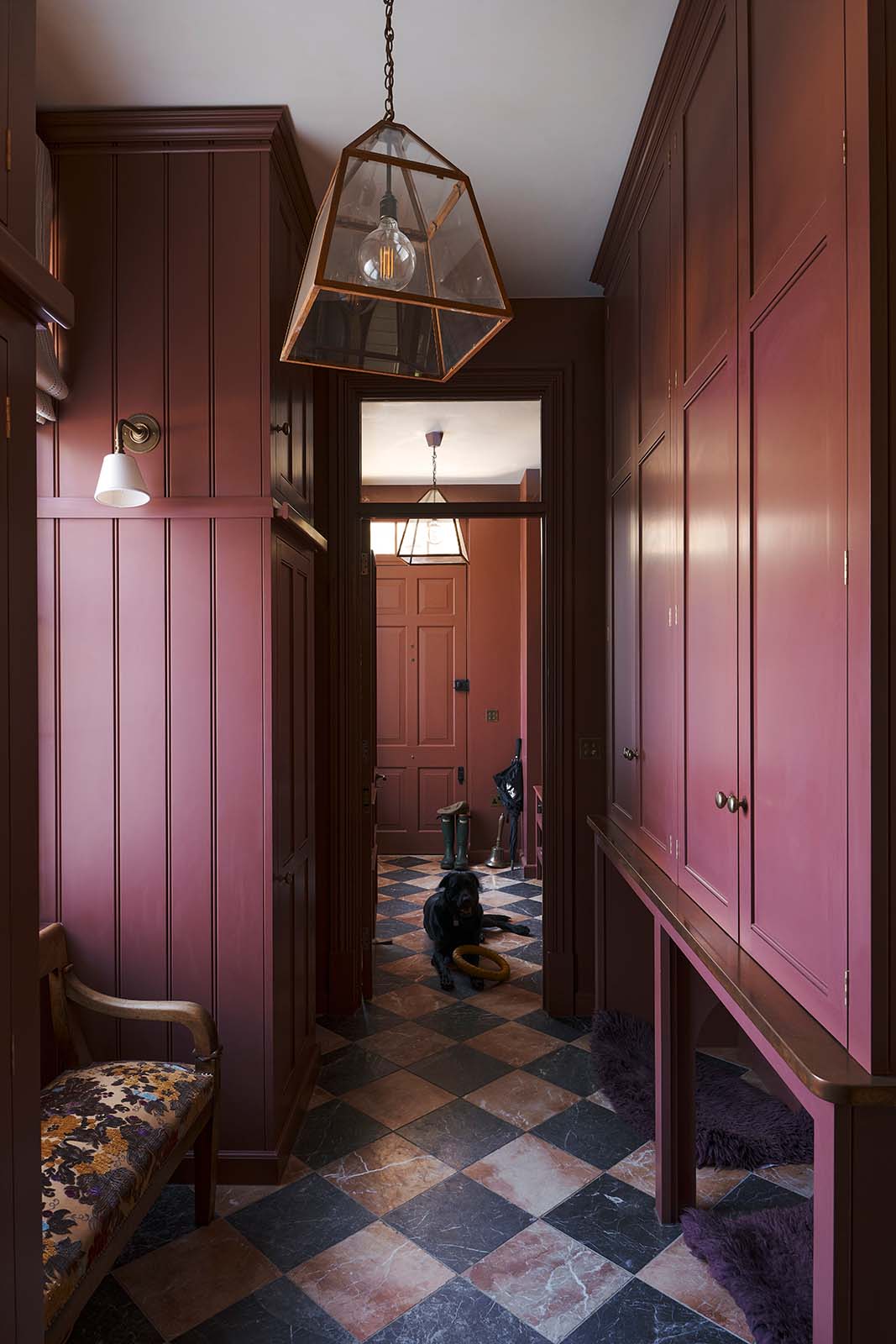 A large, refectory red hallway with full-height classic cabinets, solid oak worktop, and a black Labrador lies on the red and white marble chequered floor with a toy. The ceiling displays a glass shade with copper rims and a small white porcelain wall light.