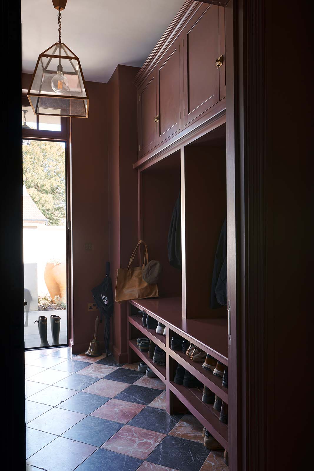 A refectory-red classic boot room features overhead cabinets, a large coat-hanging area, and open shoe shelves below. The door stands open, letting bright natural light spill into the room and across the red-and-black marble chequerboard floor.