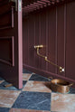 A close-up of a corner in the refectory-red boot room and hallway reveals a red-and-black marble chequered floor. Near the baseboard, a deVOL aged brass pot filler tap is installed specifically to refill the dog’s copper water bowl.
