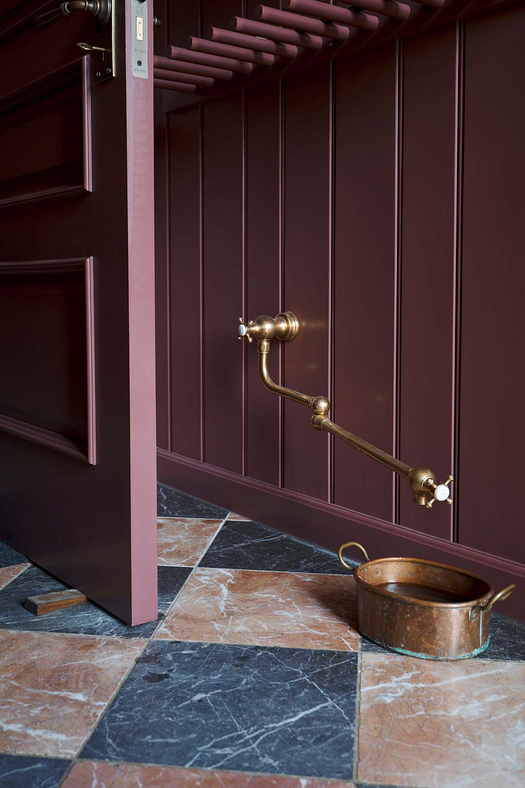 A close-up of a corner in the refectory-red boot room and hallway reveals a red-and-black marble chequered floor. Near the baseboard, a deVOL aged brass pot filler tap is installed specifically to refill the dog’s copper water bowl.