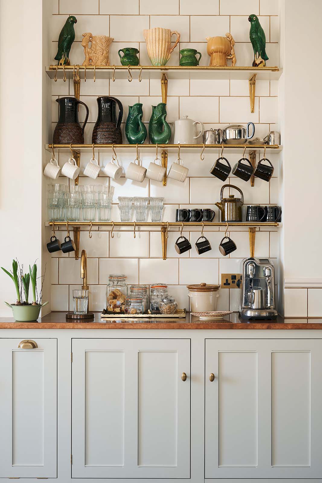 A charming personal café area features cream subway splashback tiles, three shelves with classic brass brackets, aged brass hanging rails, a Quooker Fusion mixer and instant hot tap, and a silver milk foamer. An array of simple ceramic mugs, drinking glasses, teapots, jugs, and a tray of assorted teas, coffee, and sugars completes the setup.
