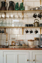 A charming personal café area features cream subway splashback tiles, three shelves with classic brass brackets, aged brass hanging rails, a Quooker Fusion mixer and instant hot tap, and a silver milk foamer. An array of simple ceramic mugs, drinking glasses, teapots, jugs, and a tray of assorted teas, coffee, and sugars completes the setup.