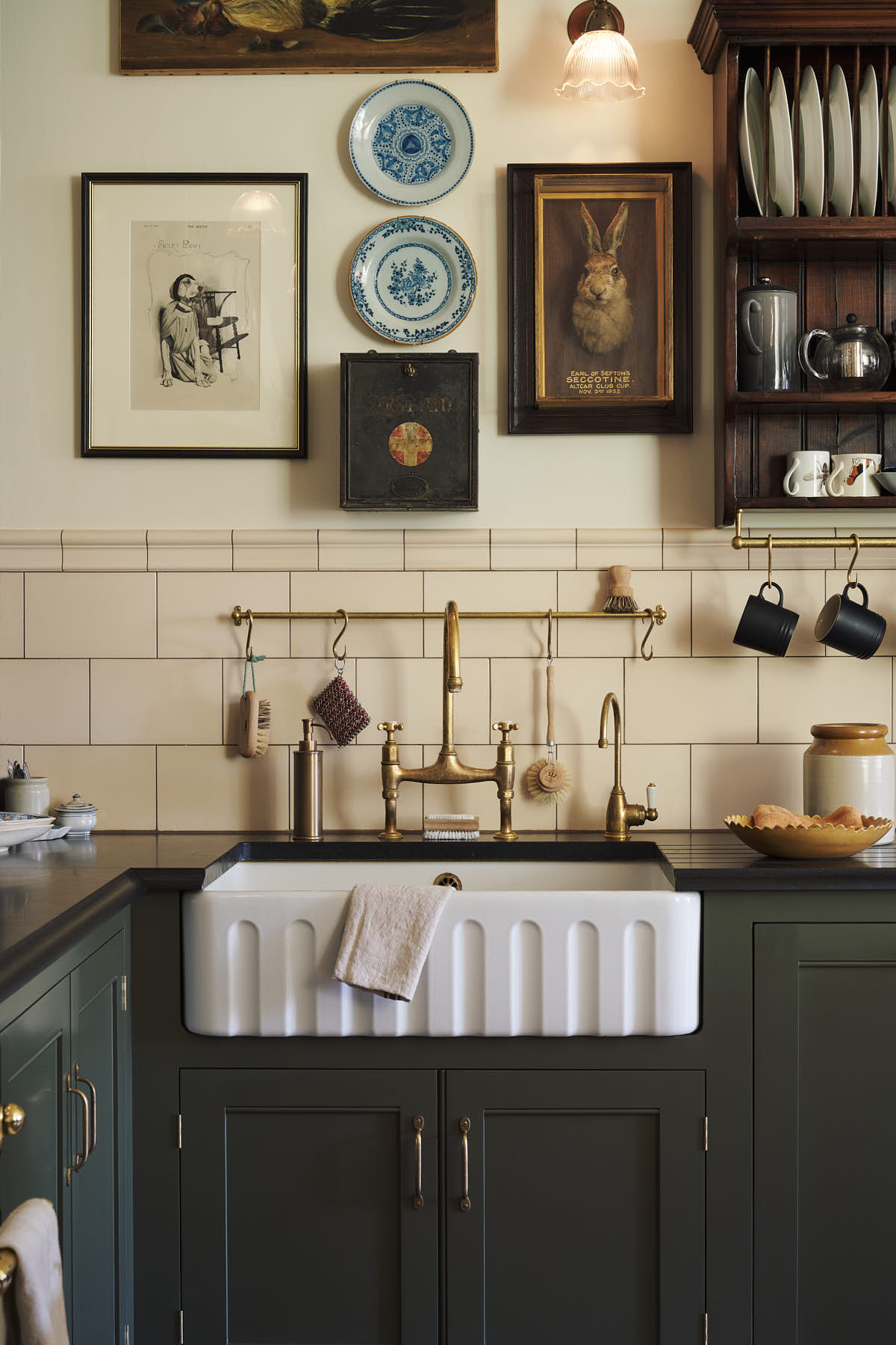 A closer look at the eccentric yet grand Bakehouse Green Classic English Kitchen features an L-shaped sink and cooker run with black granite worktop, a glazed wall cupboard, and an aged brass Ionian tap. Pridefully displaying taxidermy, artisan ceramics, and collectable antiques, it embraces a unique museum-like social kitchen space.