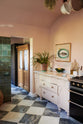 A closer look at the bright peach-coloured room reveals a chequerboard marble floor and an eclectic collection of vintage furniture, including a table and chairs, a prep table, and a decorative set of drawers. The Princelet Pink desk holds an assortment of items such as a chopping board, a ceramic bowl of lemons, a potted plant, and a gold-framed painting of a fish on a plate, giving the kitchen a wonderfully bright Mediterranean vibe.