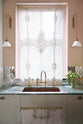 A closer view of a light pastel-blue classic kitchen’s sink run with a single copper sink with aged brass Ionian taps.  The London apartment has high ceilings, worn black painted floorboards, small decorative splashback mirrors, intricate cornices, and is styled with the customer's collection of antiques.