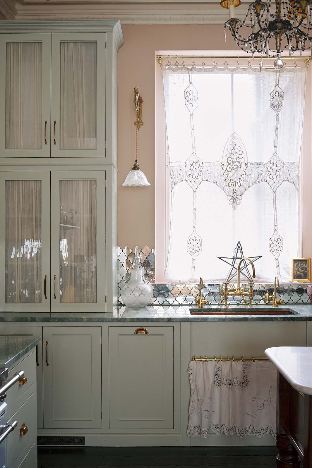 A closer view of the light pastel-blue classic kitchen’s sink run reveals a single copper sink with aged brass Ionian taps and glazed countertop cupboards. The London apartment features high ceilings, worn black-painted floorboards, small decorative splashback mirrors, intricate cornices, and is styled with the customer’s collection of antiques.