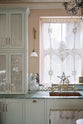 A closer view of the light pastel-blue classic kitchen’s sink run reveals a single copper sink with aged brass Ionian taps and glazed countertop cupboards. The London apartment features high ceilings, worn black-painted floorboards, small decorative splashback mirrors, intricate cornices, and is styled with the customer’s collection of antiques.