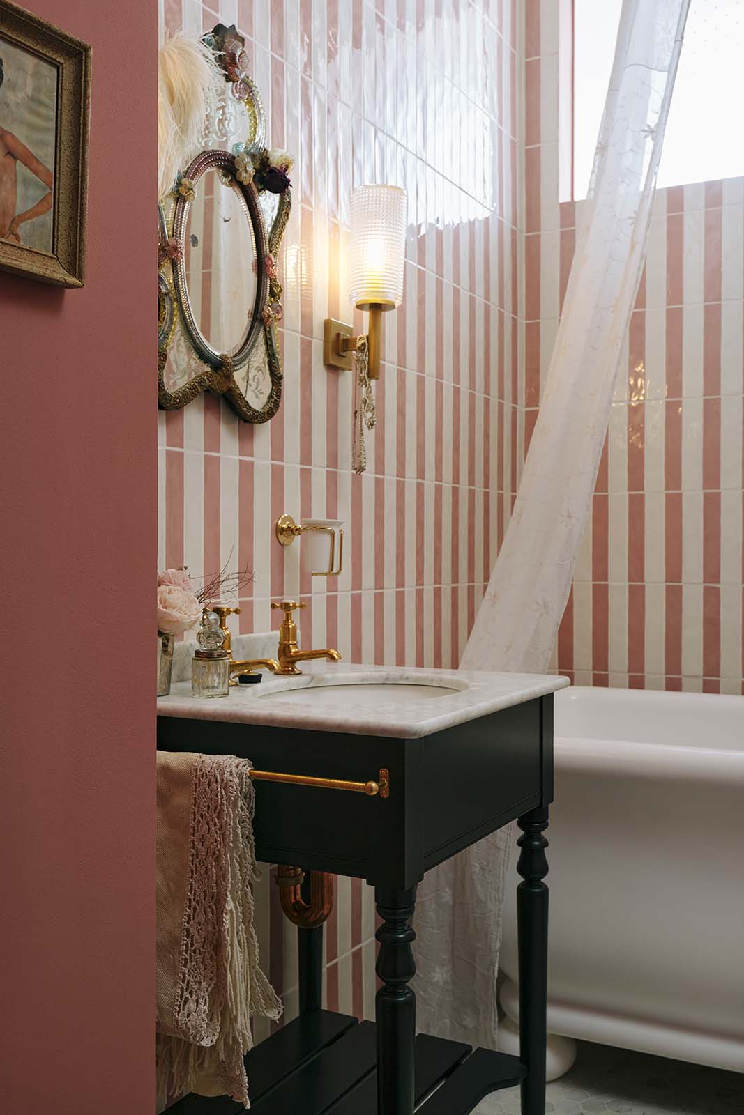 A chic bathroom with pink and white tiled walls, a white bathtub, and a small painter’s black Victorian washtand with aged brass Mayan taps. Above the sink hangs a vintage, eccentric mirror with a highly decorated frame, colourful painted florals, and a feather attached to the side.