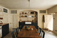 A rustic basement kitchen with sandy-coloured walls and ceiling, original Georgian detailing, cream doors and panelling with chipped paintwork and a worn stone floor. An old banquet-style table sits in the middle of the room with rows of black wooden chairs on either side. At the far side of the room, an alcove contains a small run of painted black cupboards with a mottled copper worktop, a fluted Belfast sink and an aged brass tap below shelves decorated with vintage bric-a-brac.