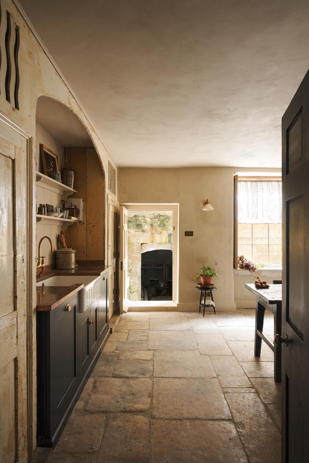 Looking through the entranceway to a rustic basement kitchen with sandy-coloured walls and ceiling, chipped paintwork and a worn stone floor. On the left is a short run of elegant black cupboards with a soft sheen and a mottled copper worktop. Ahead, light pours into the room through a large window with original wavy Georgian panes and an open doorway leading outside to a stone courtyard.