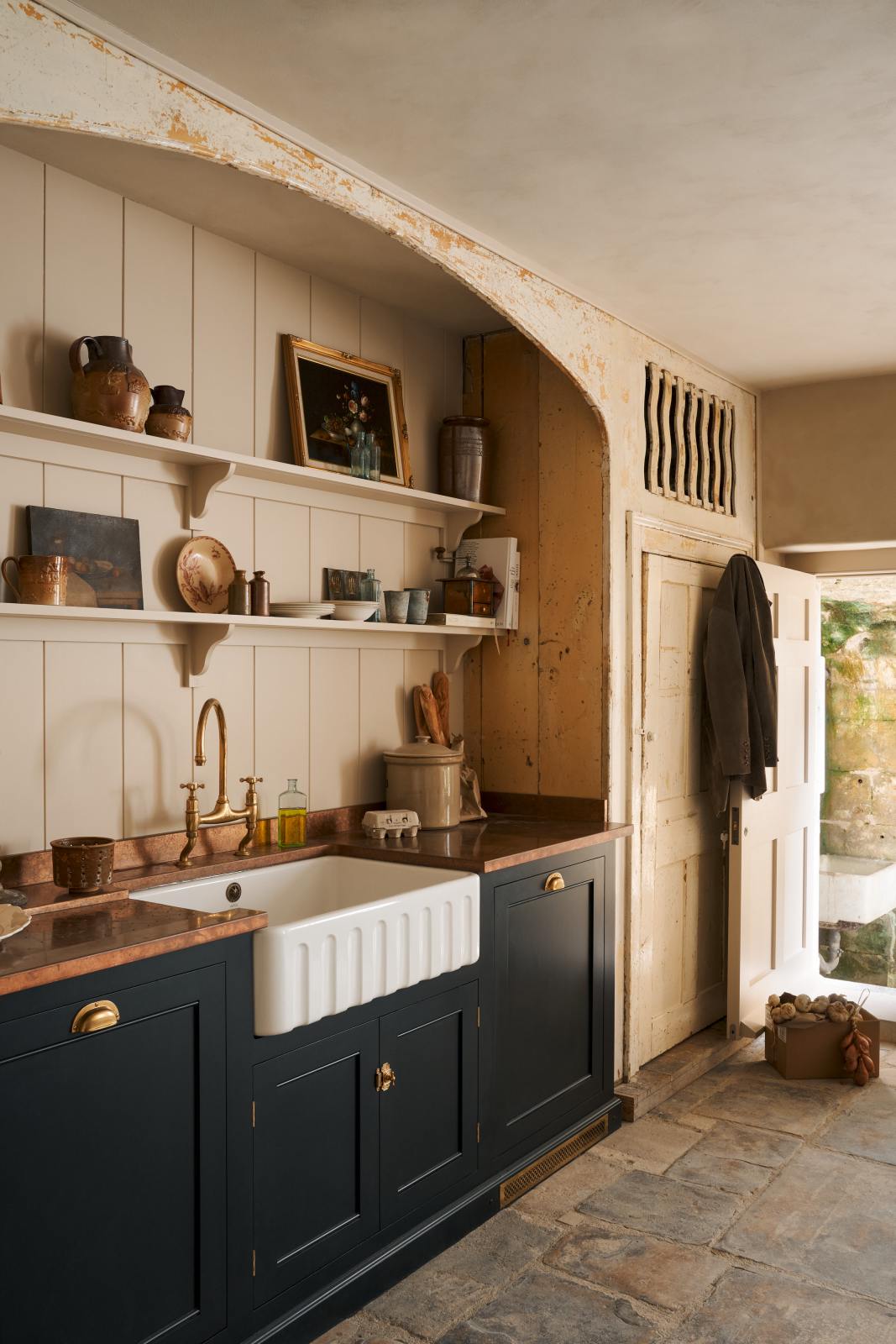 A rustic basement kitchen with sandy-coloured walls and ceiling, original Georgian detailing, cream doors and panelling with chipped paintwork and a worn stone floor. On the left is an alcove containing a small run of painted black cupboards with a mottled copper worktop, and a fluted Belfast sink below shelves decorated with vintage bric-a-brac.