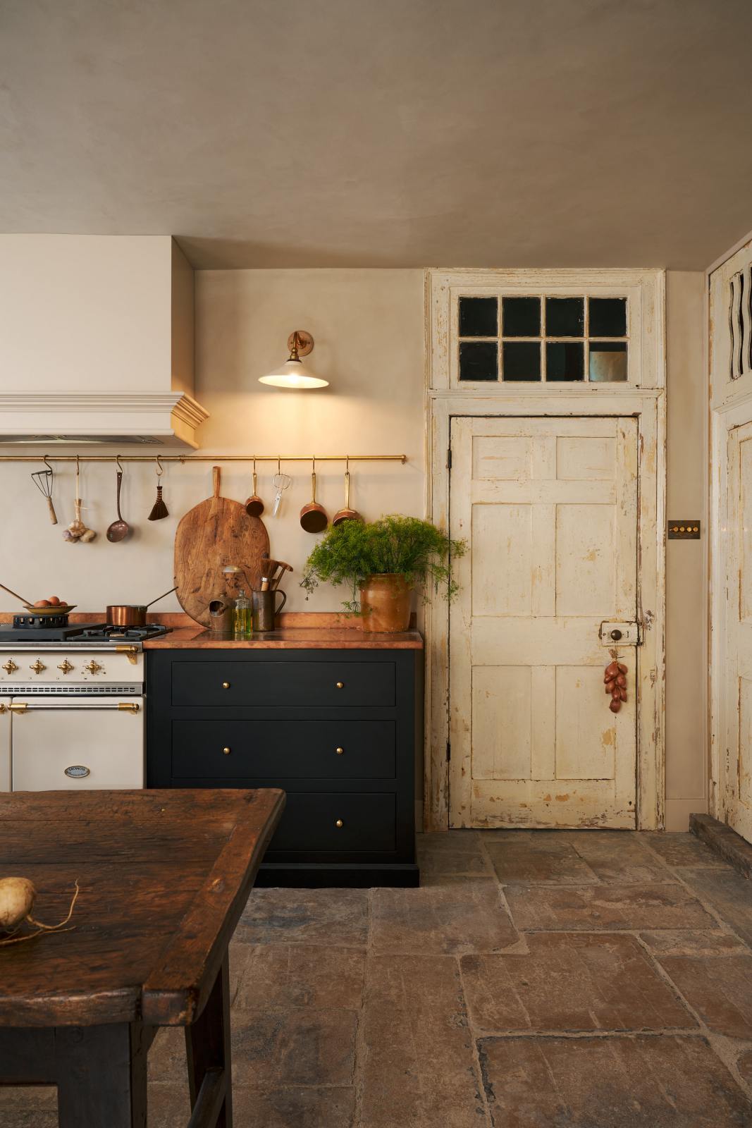 Looking through an open door of a rustic basement kitchen with sandy-coloured walls and ceiling, original Georgian detailing, cream doors and panelling with chipped paintwork and a worn stone floor. An heirloom wall light fits perfectly at home in the ancient and preserved wall.