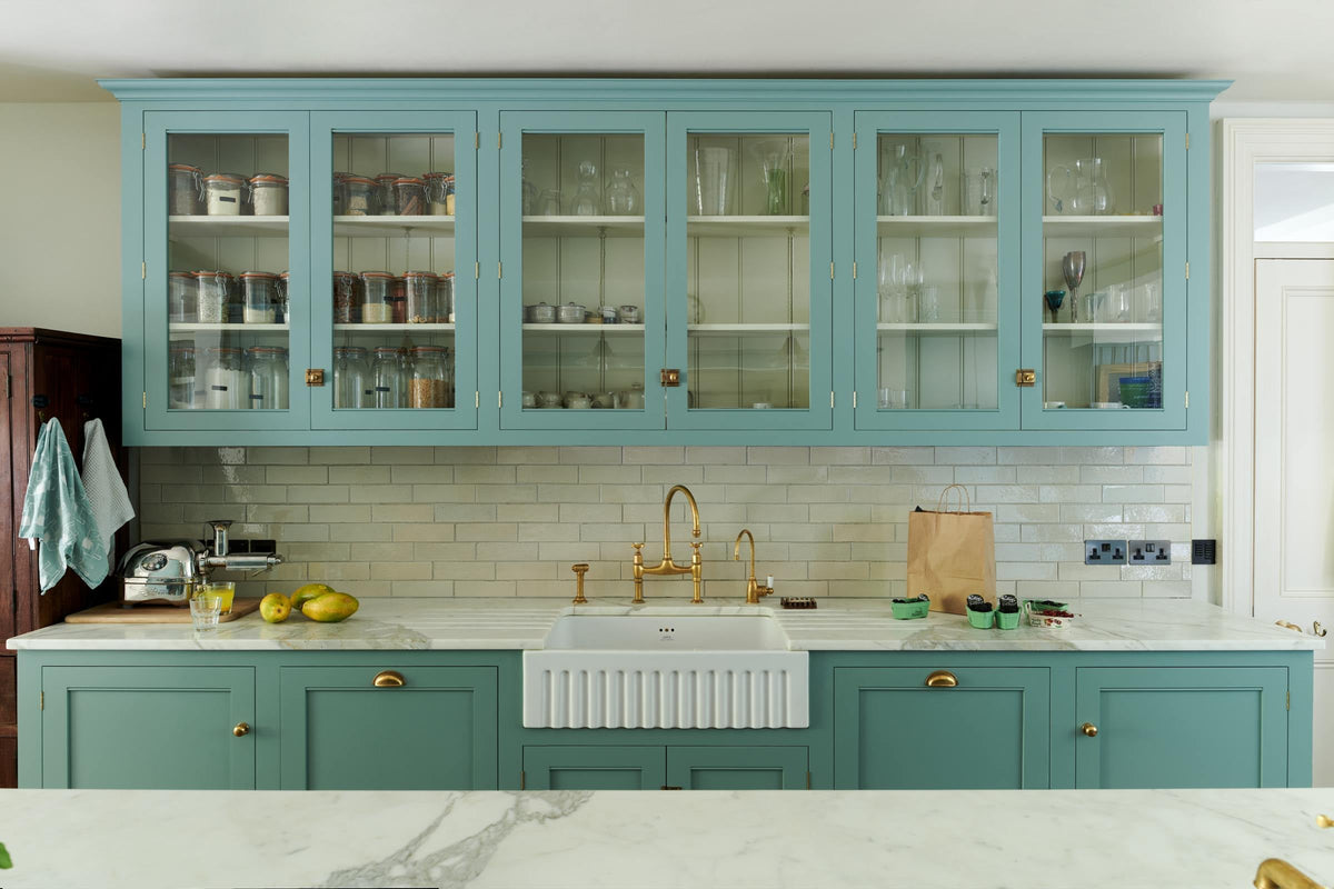 A spacious yet modest Trinity Blue classic English kitchen features a Silestone worktop sink run with a single fluted Belfast sink and aged brass Ionian tap, glazed cupboards, and a kitchen island with a copper sink—all topped off with a white porcelain splashback. Lovingly decorated with a silver juicer, papayas, live basil, berries, dried ingredients in glass jars, the room feels humble, warm, and inviting.