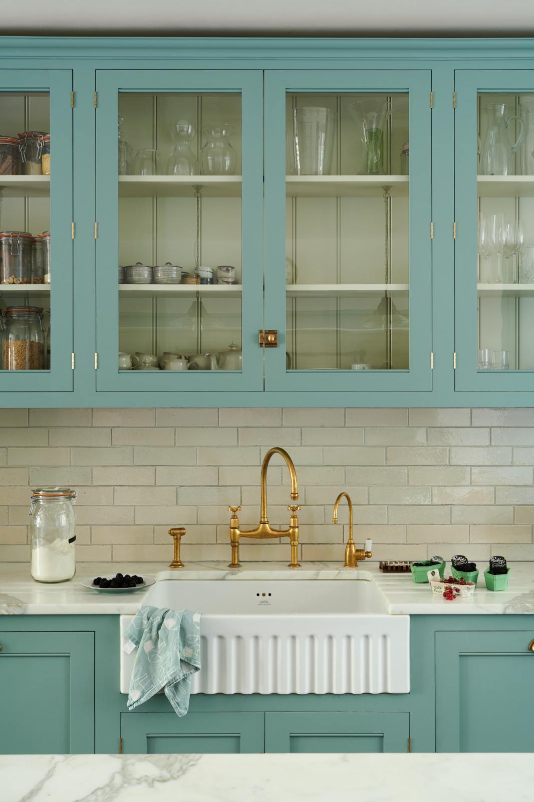 A spacious yet modest Trinity Blue classic English kitchen features a Silestone worktop sink run with a single fluted Belfast sink and aged brass Ionian tap, glazed cupboards, and a kitchen island with a copper sink—all topped off with a white porcelain splashback. Lovingly decorated with a silver juicer, papayas, live basil, berries, dried ingredients in glass jars, the room feels humble, warm, and inviting.