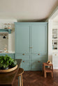 A spacious yet modest Trinity Blue classic English kitchen features a Silestone worktop cooker run, an overmantle canopy with a white porcelain splashback, a large full-height cabinet housing a fridge/freezer, a copper prep table, and deVOL wooden bum stools. Lovingly decorated with a watermelon, live basil, and dried ingredients in glass jars, the room feels humble, warm, and inviting.