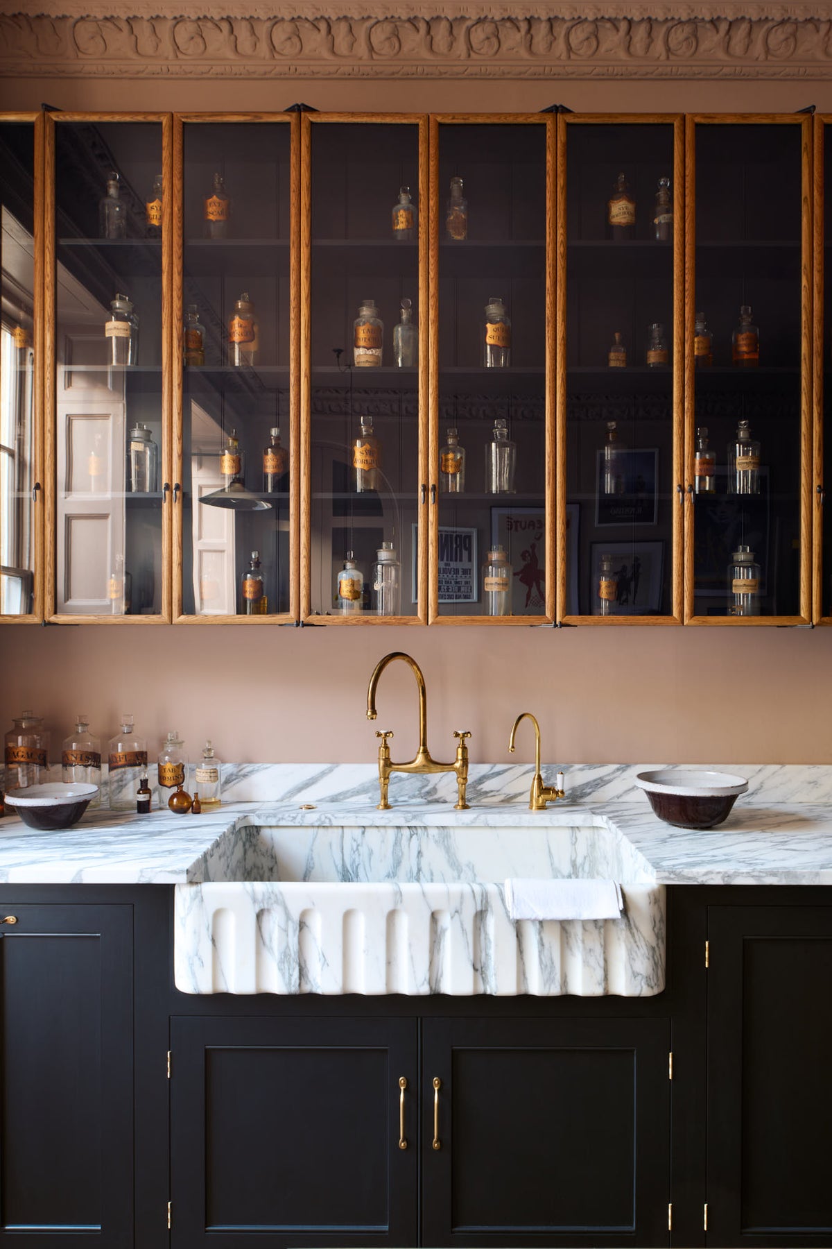 A closer look at a bold and dramatic Haberdasher’s kitchen with a sink and cooker run, an Arabescato worktop and single fluted sink, an aged brass Ionian tap, and a glazed wall cupboard. Adorned with artisan ceramics, labelled glass jars, and a quirky framed illustration, creating a local bristo bar in a Manhattan space.