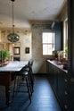 An elegant classic kitchen includes a sink run with a copper sink and aged brass Ionian tap, an aged copper worktop, wall cupboards, a marble worktop heirloom dairy table, a breakfast nook, and tall black chairs. The tall-ceilinged room has a rustic wash finish, with green draping down the walls, resembling overgrown ancient ruins. Beyond the archways leading to the living room is a striking black fireplace, a black-framed mirror, and a woven chandelier.