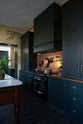 A closer look at an elegant classic kitchen’s cooker run reveals an aged copper worktop, a shaker canopy, counter cabinets, and a marble worktop heirloom dairy table. The pantry-blue counter cabinets and canopy stand tall, and in the centre is a black gas cooker, copper pans, and fresh chillies hanging from aged brass rails, with a large aged copper splashback behind.
