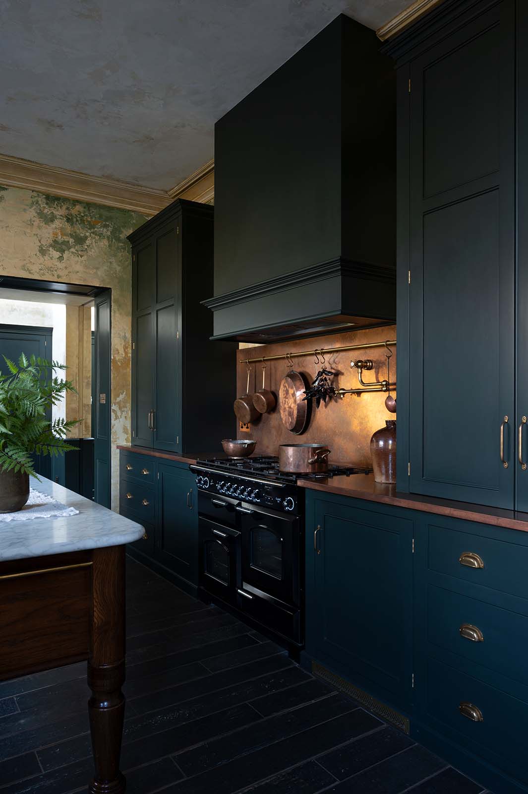 A closer look at an elegant classic kitchen’s cooker run reveals an aged copper worktop, a shaker canopy, counter cabinets, and a marble worktop heirloom dairy table. The pantry-blue counter cabinets and canopy stand tall, and in the centre is a black gas cooker, copper pans, and fresh chillies hanging from aged brass rails, with a large aged copper splashback behind.