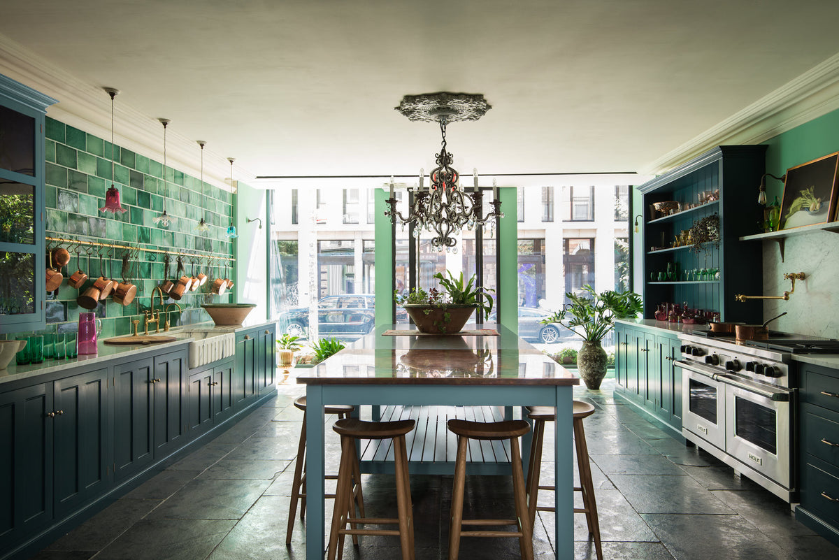 A stunning, Bond Street blue kitchen features a sink and cooker run with Arabescato marble worktop, a bespoke prep table, a large wall cupboard, and a bank of open pantry larder. The room features a stunning mix of colours and character, accentuated by elegant antiques such as a crystal chandelier, colourful glass pendant lights, aged brass rails with copper pans, an aged brass pot filler, and deVOL Emerald green London splashback tiles.