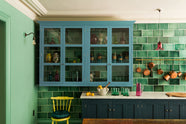 A stunning, Bond Street blue kitchen features a sink run with Arabescato marble worktop, a bespoke prep table, and a large wall cupboard. The room features a stunning mix of colours and character, accentuated by elegant antiques such as artisan ceramics, colourful glass pendant lights, aged brass rails with copper pans, and deVOL Emerald green London splashback tiles.