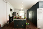 This grand bakehouse green classic English kitchen has large full-height pantry cupboards, oven cabinets, fridge/freezer cupboards, a long kitchen island with a double sink, an aged brass Ionian tap, and a curiosity cupboard. Adorned with white and blush pink hydrangeas, romanesco cauliflower, potted plants, still life paintings, a candelabra, and homemade jams, the room feels homey and welcoming despite its large and decorative appearance.