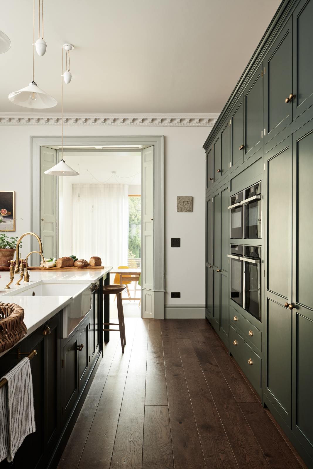 Looking between a large full-height pantry cupboard, oven cabinets, fridge/freezer cupboard, and a long kitchen island with a double sink, an aged brass Ionian tap in the grand bakehouse green classic English kitchen. Adorned with loaves of bread, leeks, potted plants, and still life paintings; the room feels homey and welcoming despite its large and decorative appearance.